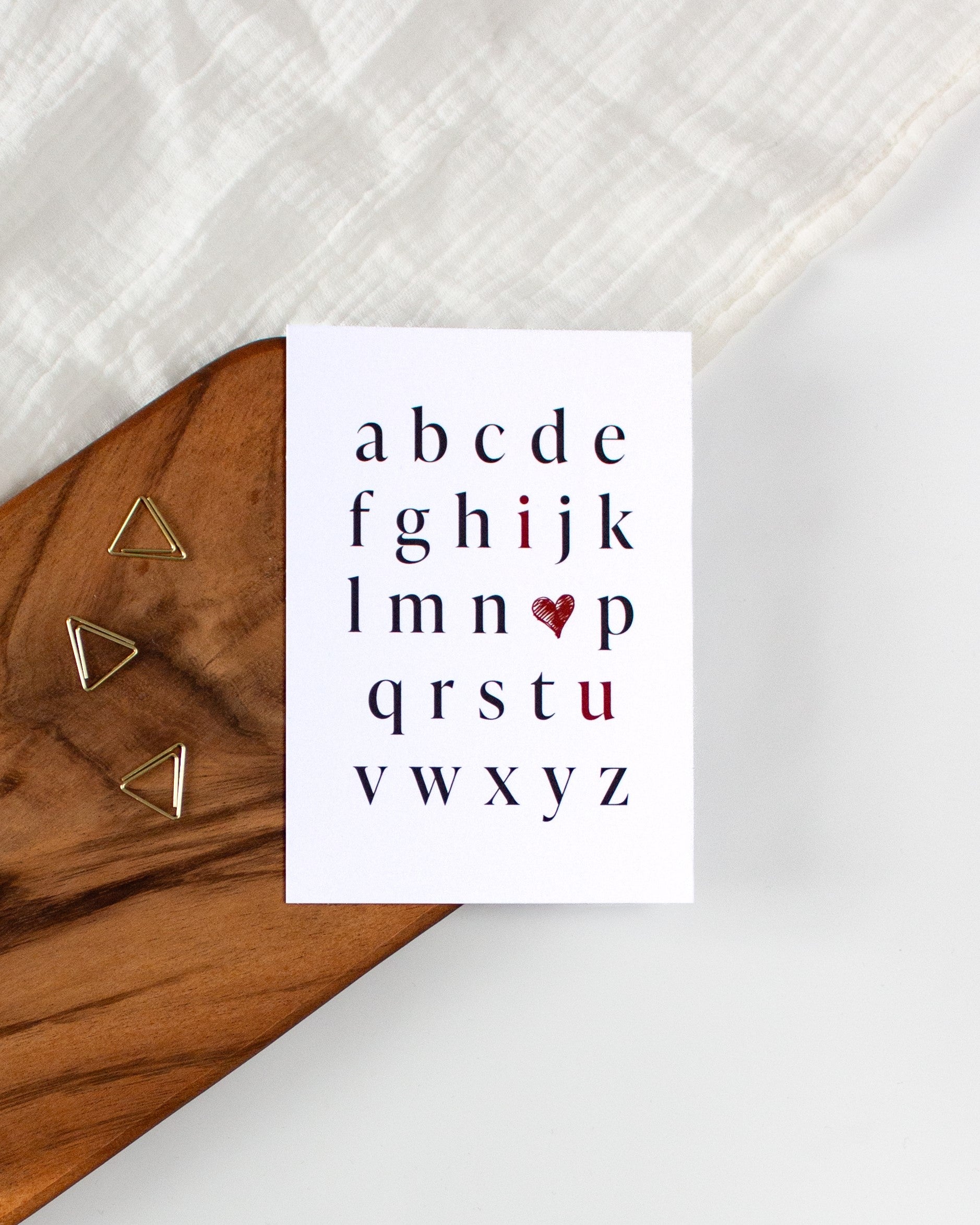 A postcard laying on a wooden board with some golden triangle paperclips and a white cloth in the background. The postcard design shows the alphabet in mostly black letters. The letters "i" and "u" are red. In place of the letter "o" there is a red heart. The letters are arranged in a way that the red letters and heart read "I love you".
