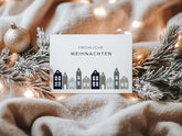 Minimalistische Weihnachtskarte im Querformat mit dem Gruß "Fröhliche Weihnachten" und einer Reihe stilisierter Dorfhäuser in gedeckten Farben, präsentiert auf einer flauschigen Decke mit schneebedecktem Tannenzweig, einer glänzenden Weihnachtskugel und Lichterketten für eine gemütliche Winteratmosphäre.