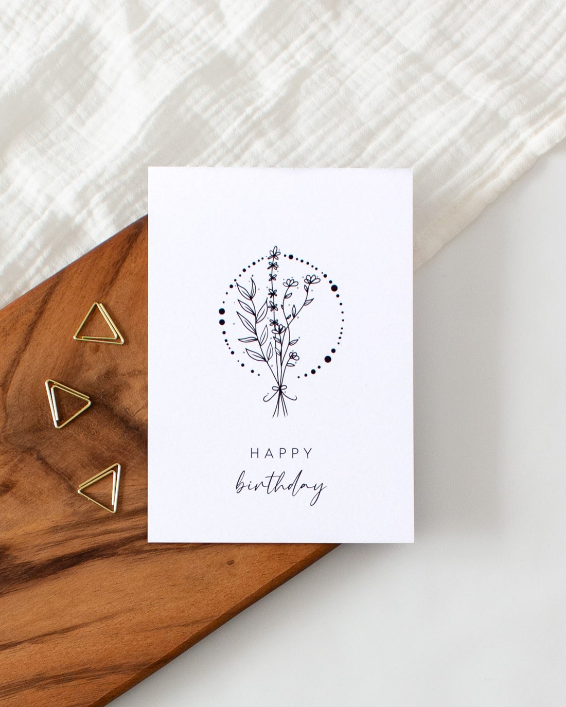 A postcard laying on a wooden board with some golden triangle paperclips and a white cloth in the background. The postcard design shows a lineart drawing of some flowers and branches tied together, with a circle consisting of dots of varying sizes around them. Below that are two lines of text saying &quot;Happy Birthday&quot;.