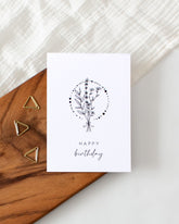 A postcard laying on a wooden board with some golden triangle paperclips and a white cloth in the background. The postcard design shows a lineart drawing of some flowers and branches tied together, with a circle consisting of dots of varying sizes around them. Below that are two lines of text saying "Happy Birthday".