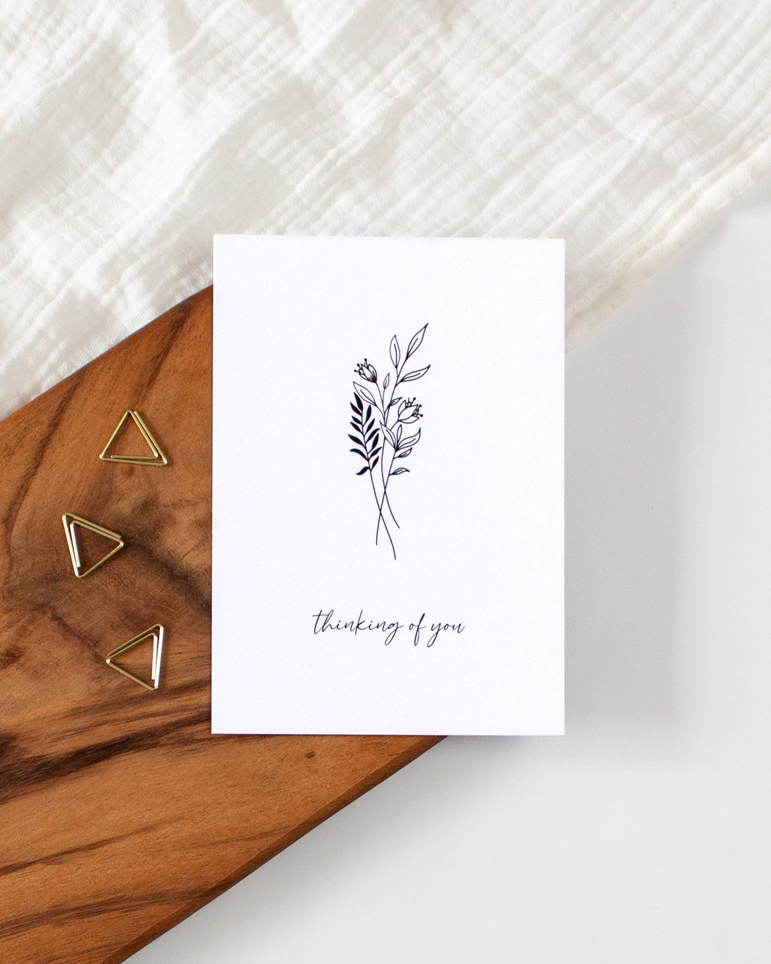 A postcard laying on a wooden board with some golden triangle paperclips and a white cloth in the background. The postcard design shows a line art drawing of some flowers and leaves with a line of cursive writing below, that says &quot;thinking of you&quot;.