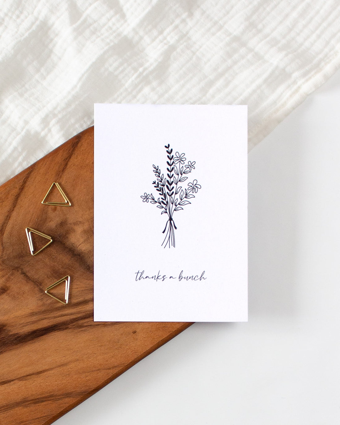 A postcard laying on a wooden board with some golden triangle paperclips and a white cloth in the background. The postcard design shows a line art drawing of a bouquet of some flowers and branches, with a line of cursive writing below, that says &quot;thanks a bunch&quot;.
