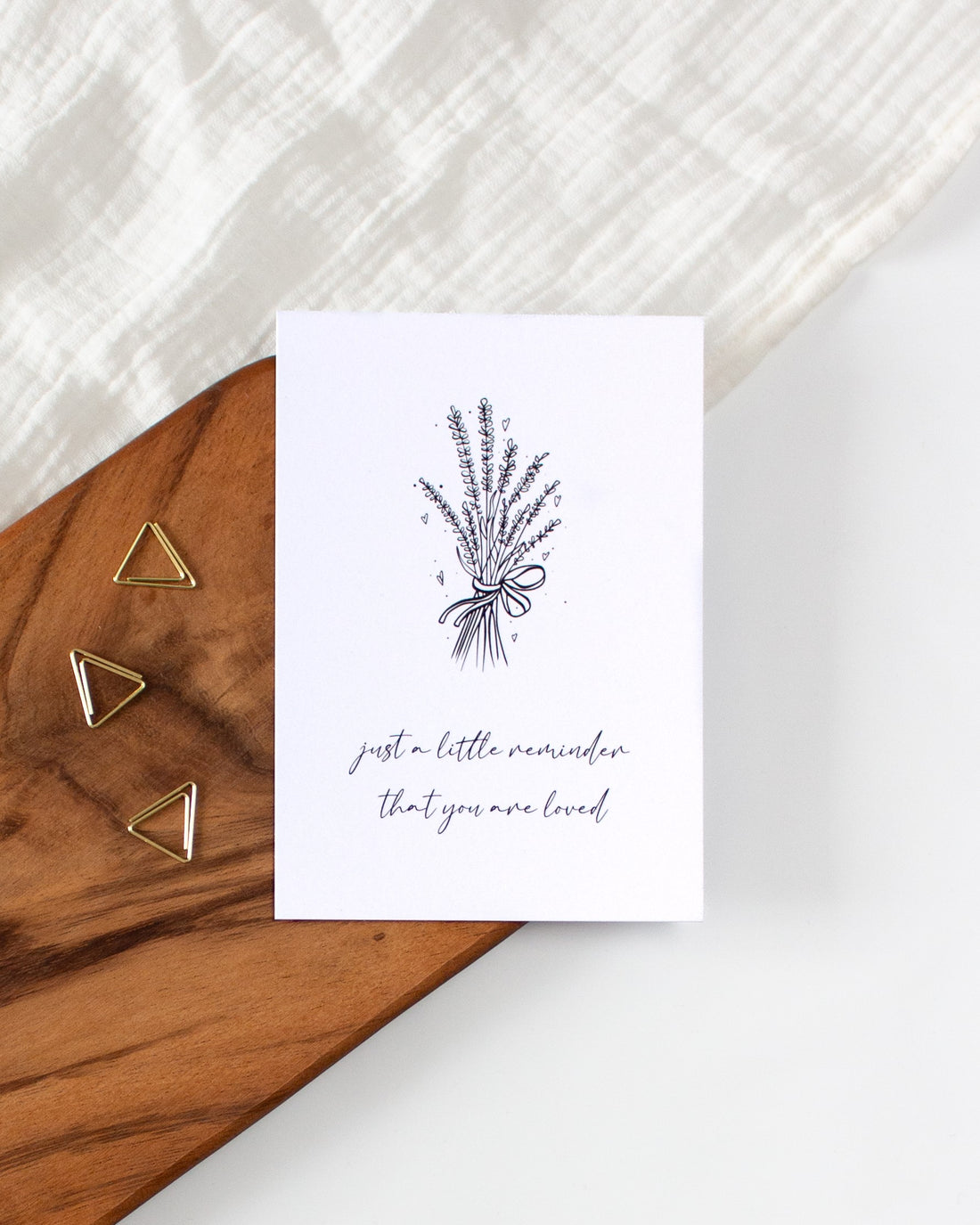 A postcard laying on a wooden board with some golden triangle paperclips and a white cloth in the background. The postcard design has a line art drawing of a flower bouquet with little hearts. Below that are two lines of cursive writing saying &quot;Just a little reminder that you are loved&quot;.