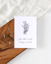 A postcard laying on a wooden board with some golden triangle paperclips and a white cloth in the background. The postcard design has a line art drawing of a flower bouquet with little hearts. Below that are two lines of cursive writing saying "Just a little reminder that you are loved".