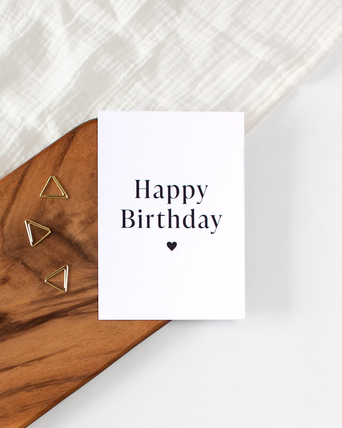 A postcard laying on a wooden board with golden triangle paperclips and a white cloth in the background. The postcard design consists of two big lines of text saying &quot;Happy Birthday&quot; and a small heart below them.