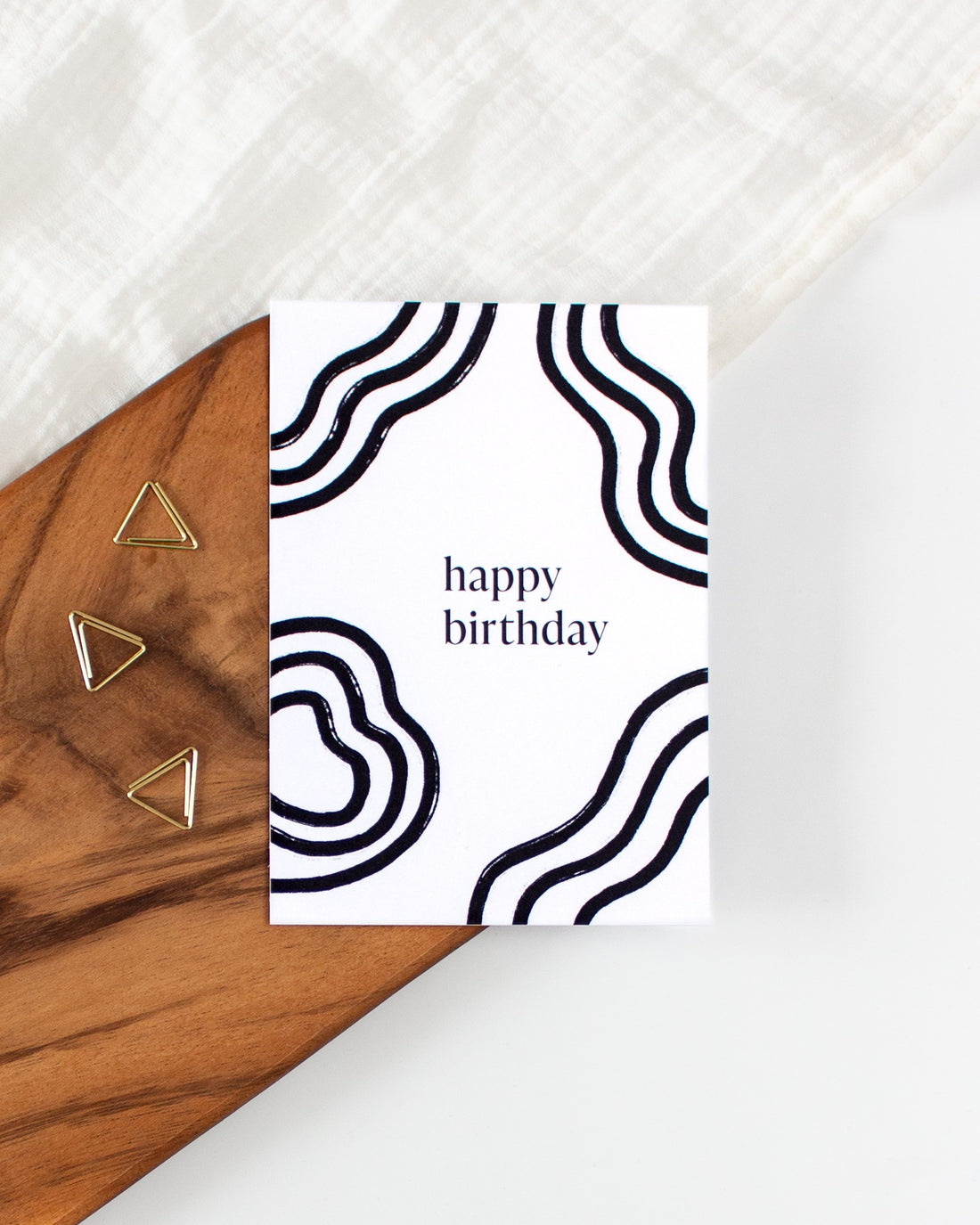 A postcard laying on a wooden board with some golden triangle paperclips and a white cloth in the background. The postcard design consists of bold black wavy lines and text saying &quot;happy birthday&quot;.