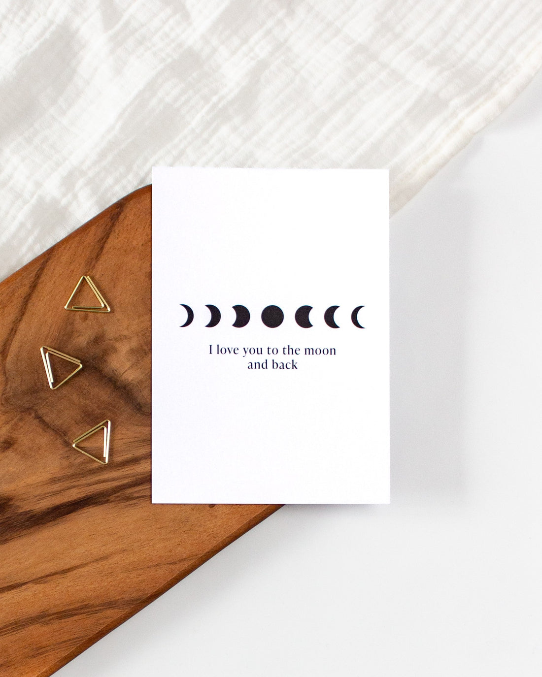 A white postcard laying on a wooden board with some golden triangle paperclips and a white cloth in the background. The postcard design shows simple black symbols of moon phases arranged in a horizontal line with some text below them saying &quot;I love you to the moon and back&quot;.