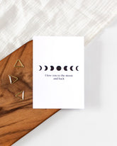 A white postcard laying on a wooden board with some golden triangle paperclips and a white cloth in the background. The postcard design shows simple black symbols of moon phases arranged in a horizontal line with some text below them saying "I love you to the moon and back".