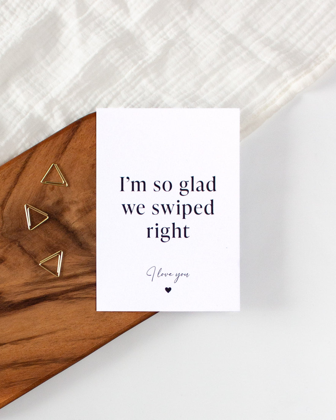 A white postcard laying on a wooden board with some golden triangle paperclips and a white cloth in the background. The postcard design reads &quot;I&