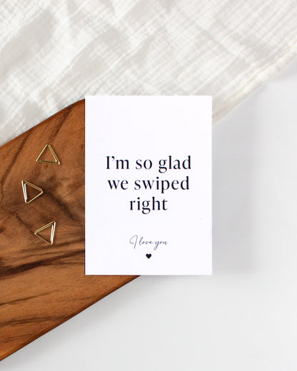 A white postcard laying on a wooden board with some golden triangle paperclips and a white cloth in the background. The postcard design reads &quot;I&