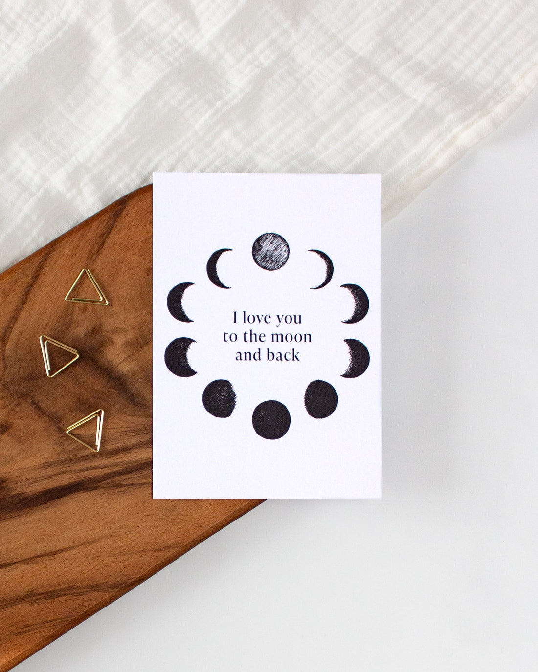 A white postcard laying on a wooden board with some golden triangle paperclips and a white cloth in the background.. The postcard design shows black sketches of moon phases arranged in a circle with some text in the middle saying &quot;I love you to the moon and back&quot;.