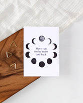 A white postcard laying on a wooden board with some golden triangle paperclips and a white cloth in the background.. The postcard design shows black sketches of moon phases arranged in a circle with some text in the middle saying "I love you to the moon and back".