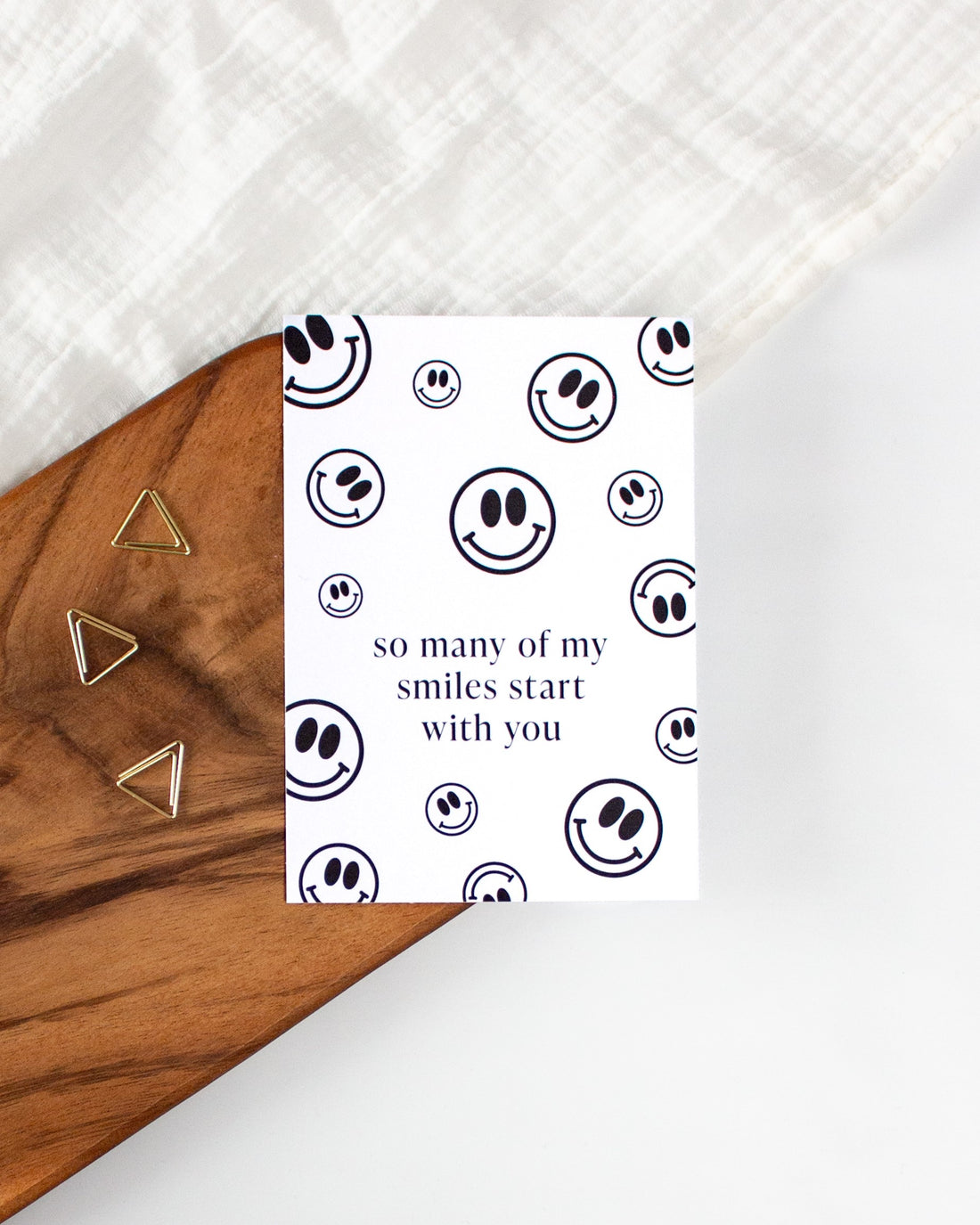 A white postcard laying on a wooden board with some golden triangle paperclips and a white cloth in the background. The postcard is covered in black smiley faces of varying sizes with some text in the middle saying &quot;so many of my smiles start with you&quot;.