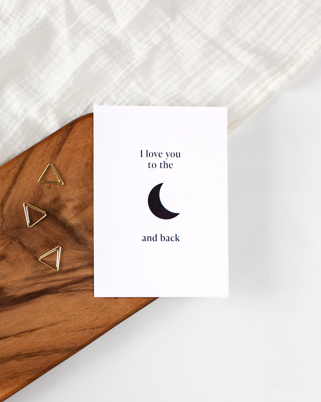 A white postcard laying on a wooden board with some golden triangle paperclips and a white cloth in the background. The postcard design shows a simple black moon symbol in the middle with text above it saying &quot;I love you to the&quot; and text below it saying &quot;and back&quot;.