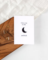 A white postcard laying on a wooden board with some golden triangle paperclips and a white cloth in the background. The postcard design shows a simple black moon symbol in the middle with text above it saying "I love you to the" and text below it saying "and back".