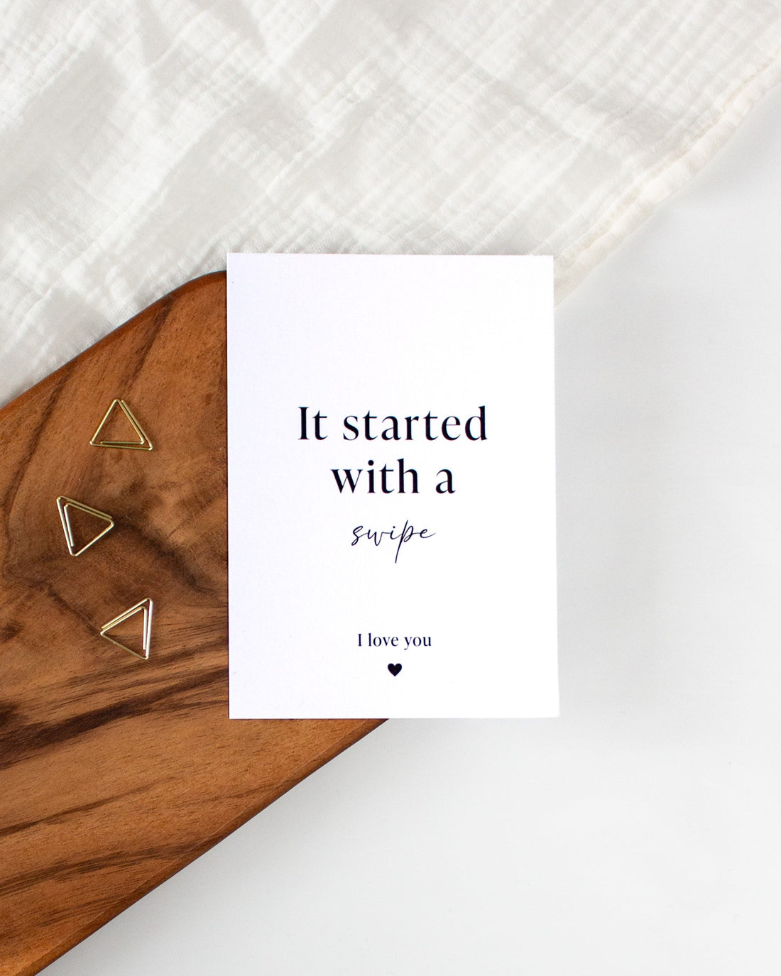 A white postcard with black writing laying on a wooden board with some golden triangle paperclips and a white cloth in the background. The postcard reads &quot;It started with a swipe&quot; in the middle and &quot;I love you&quot; in smaller text with a small heart in the bottom of the card.