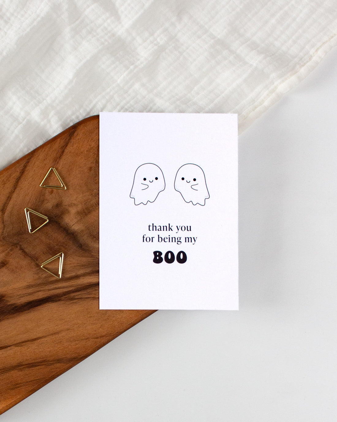 A postcard laying on a wooden board with some golden triangle paperclips and a white cloth in the background. The postcard design shows two adorable ghost drawings and some text saying &quot;thank you for being my boo&quot;.
