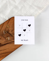 A white postcard laying on a wooden board with some golden triangle paperclips and a white cloth in the background. The postcard design shows a game of tic-tac-toe being won with heart symbols. Text saying "you won my heart" is split up with the first two words being above the game and the rest below it.