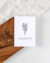 A postcard laying on a wooden board with some golden triangle paperclips and a white cloth in the background. The postcard design consists of a line art drawing of a bunch of flowers tied together and some cursive writing saying "Happy Birthday" below that.