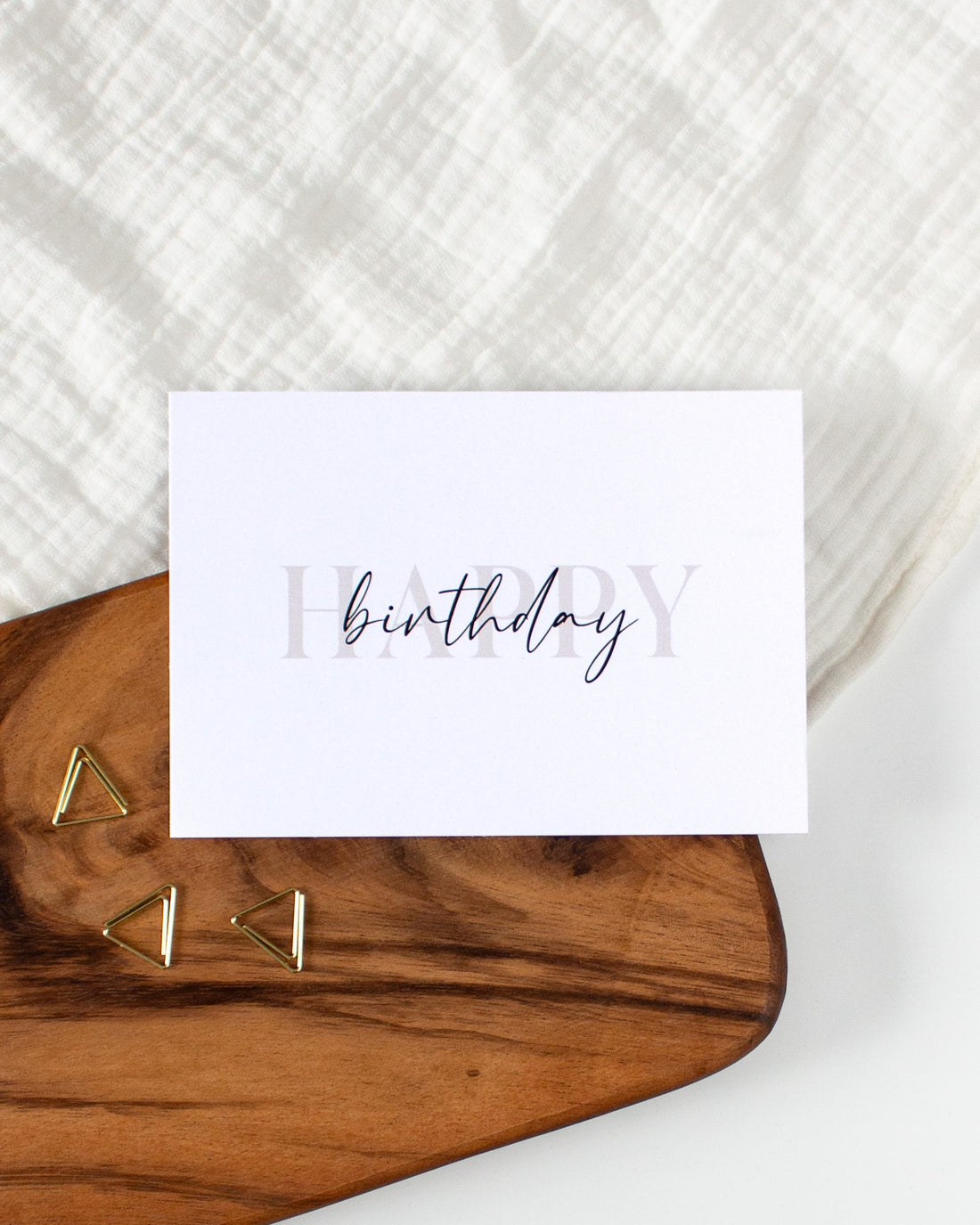 A postcard laying on a wooden board with some golden triangle paperclips and a white cloth in the background. The postcard design consists of bold beige letters saying &quot;Happy&quot; with black cursive writing on top saying &quot;birthday&quot;.