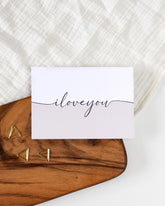 A postcard laying on a wooden board with some golden triangle paperclips and a white cloth in the background. The postcard design reads "I love you" in black cursive writing, that splits the card into two halves. The top half is white and the bottom half is beige.