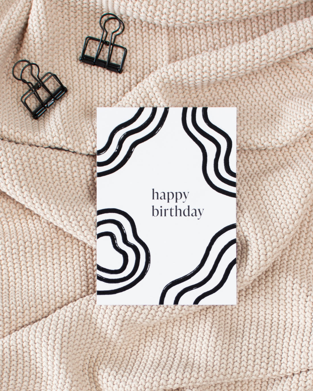 A postcard laying on a beige knitted blanket with some black binder clips. The postcard design consists of bold black wavy lines and text saying &quot;happy birthday&quot;.