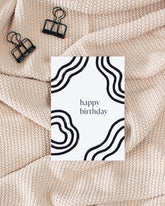 A postcard laying on a beige knitted blanket with some black binder clips. The postcard design consists of bold black wavy lines and text saying "happy birthday".