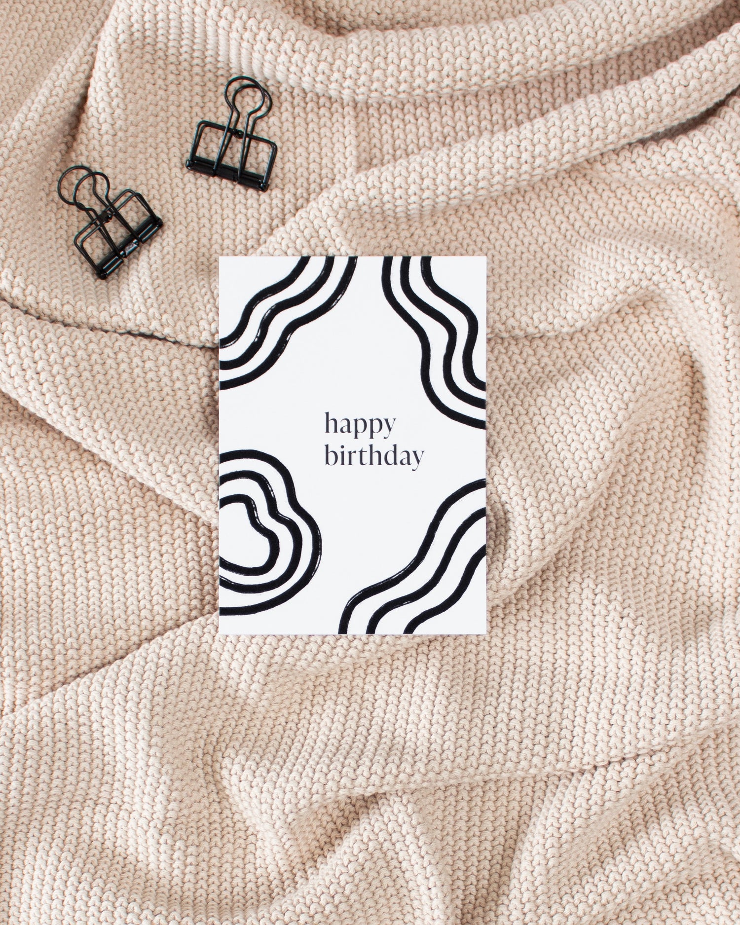 weiße Happy Birthday Geburtstagskarte mit schwarzem Linien-Design auf beiger Strick-Decke mit schwarzen Foldback-Klammern