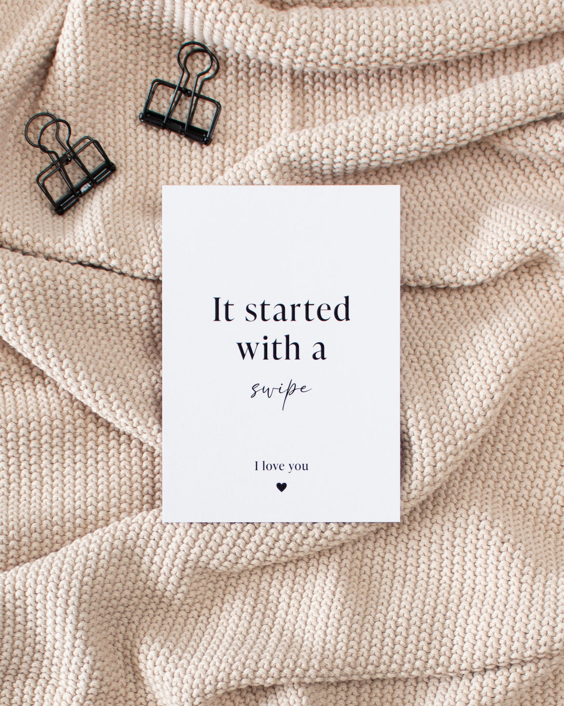 A white postcard with black writing laying on a beige knitted blanket with some black binder clips. The postcard reads &quot;It started with a swipe&quot; in the middle and &quot;I love you&quot; in smaller text with a small heart in the bottom of the card.
