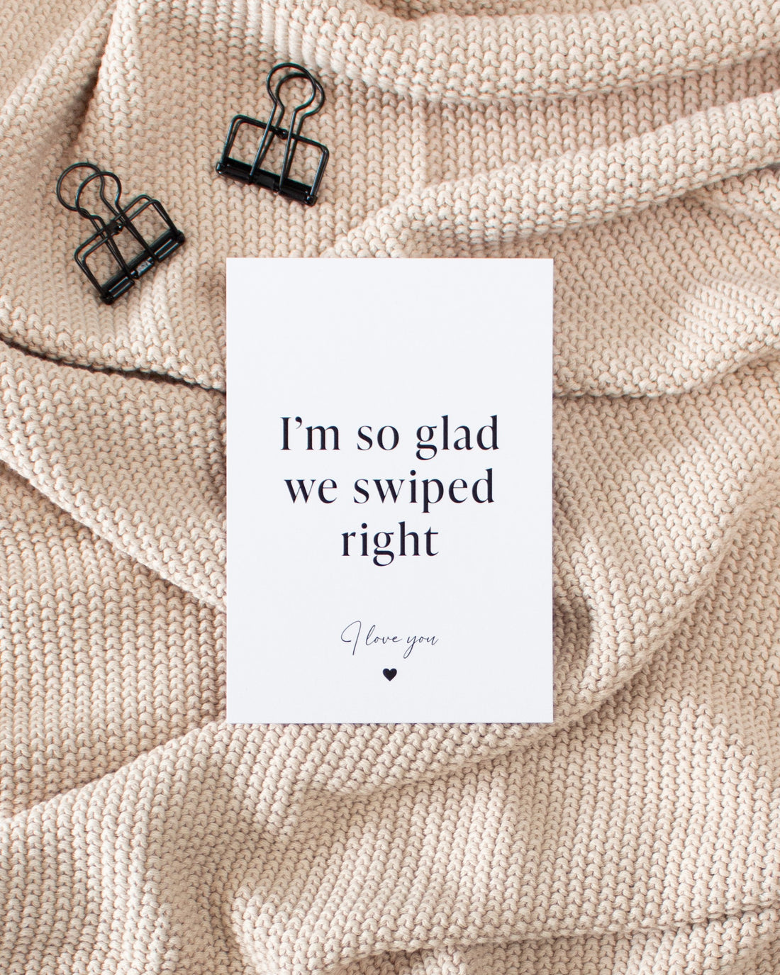 A white postcard laying on a beige knitted blanket with some black binder clips. The postcard design reads &quot;I&