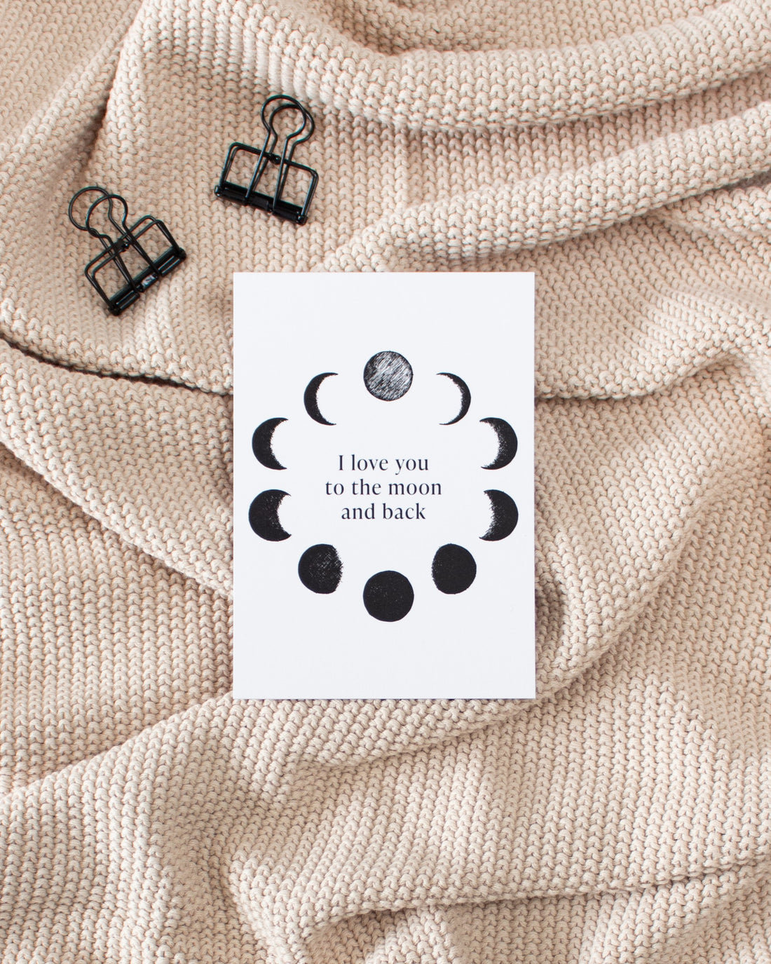 A white postcard laying on a beige knitted blanket with some black binder clips. The postcard design shows black sketches of moon phases arranged in a circle with some text in the middle saying &quot;I love you to the moon and back&quot;.