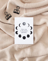 A white postcard laying on a beige knitted blanket with some black binder clips. The postcard design shows black sketches of moon phases arranged in a circle with some text in the middle saying "I love you to the moon and back".