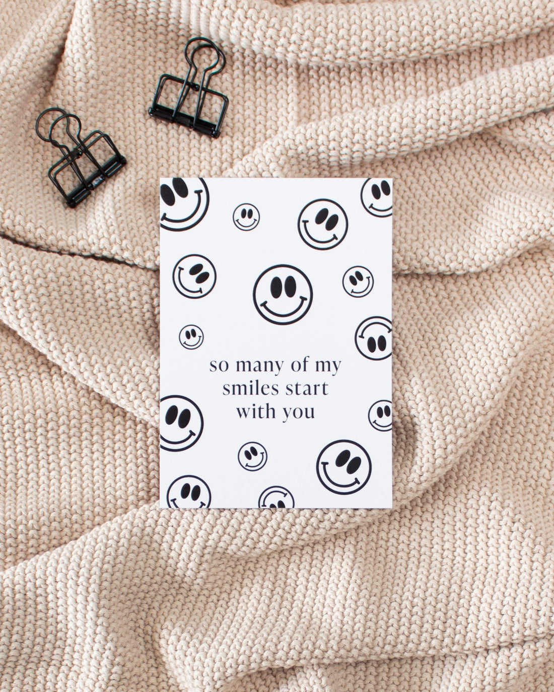 A white postcard laying on a beige knitted blanket with some black binder clips. The postcard is covered in black smiley faces of varying sizes with some text in the middle saying &quot;so many of my smiles start with you&quot;.