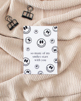 A white postcard laying on a beige knitted blanket with some black binder clips. The postcard is covered in black smiley faces of varying sizes with some text in the middle saying "so many of my smiles start with you".