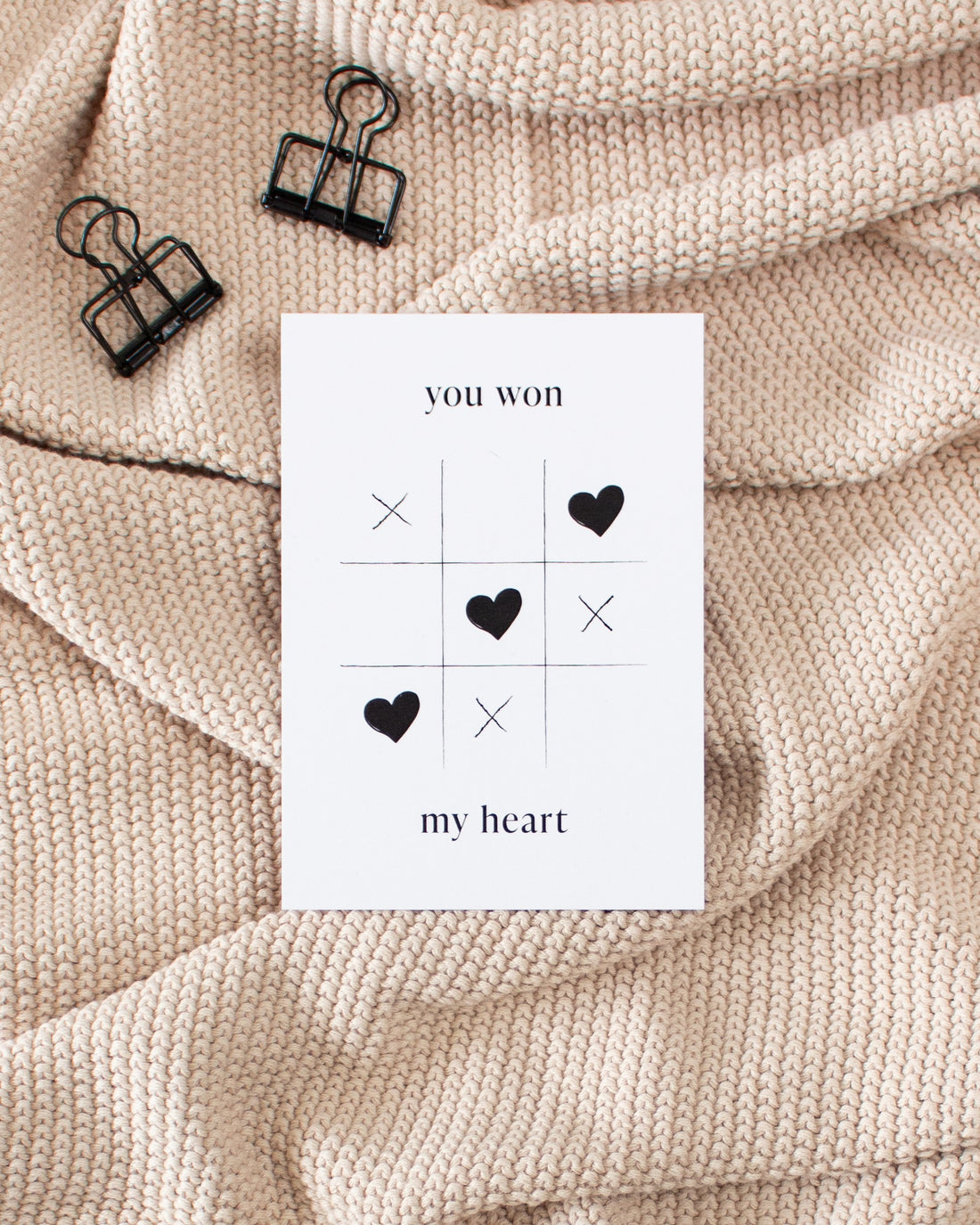 A white postcard laying on a beige knitted blanket with some black binder clips. The postcard design shows a game of tic-tac-toe being won with heart symbols. Text saying &quot;you won my heart&quot; is split up with the first two words being above the game and the rest below it.