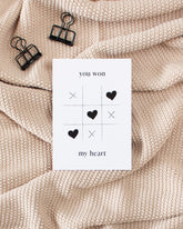 A white postcard laying on a beige knitted blanket with some black binder clips. The postcard design shows a game of tic-tac-toe being won with heart symbols. Text saying "you won my heart" is split up with the first two words being above the game and the rest below it.