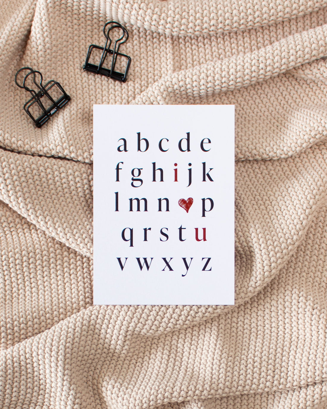 A postcard laying on a beige knitted blanket with some black binder clips. The postcard design shows the alphabet in mostly black letters. The letters &quot;i&quot; and &quot;u&quot; are red. In place of the letter &quot;o&quot; there is a red heart. The letters are arranged in a way that the red letters and heart read &quot;I love you&quot;.