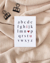 A postcard laying on a beige knitted blanket with some black binder clips. The postcard design shows the alphabet in mostly black letters. The letters "i" and "u" are red. In place of the letter "o" there is a red heart. The letters are arranged in a way that the red letters and heart read "I love you".