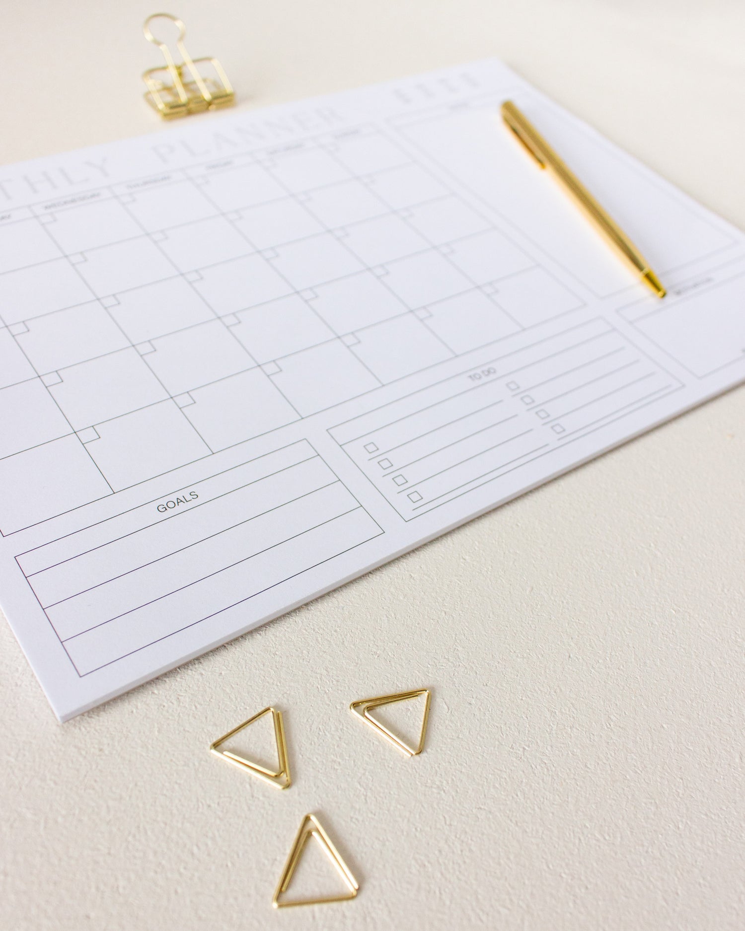 undatierter Monatsplaner Notizblock mit goldenen Dreiecks-Büroklammern, goldenem Kugelschreiber und goldener Foldback-Klammer auf hell-beigem Hintergrund