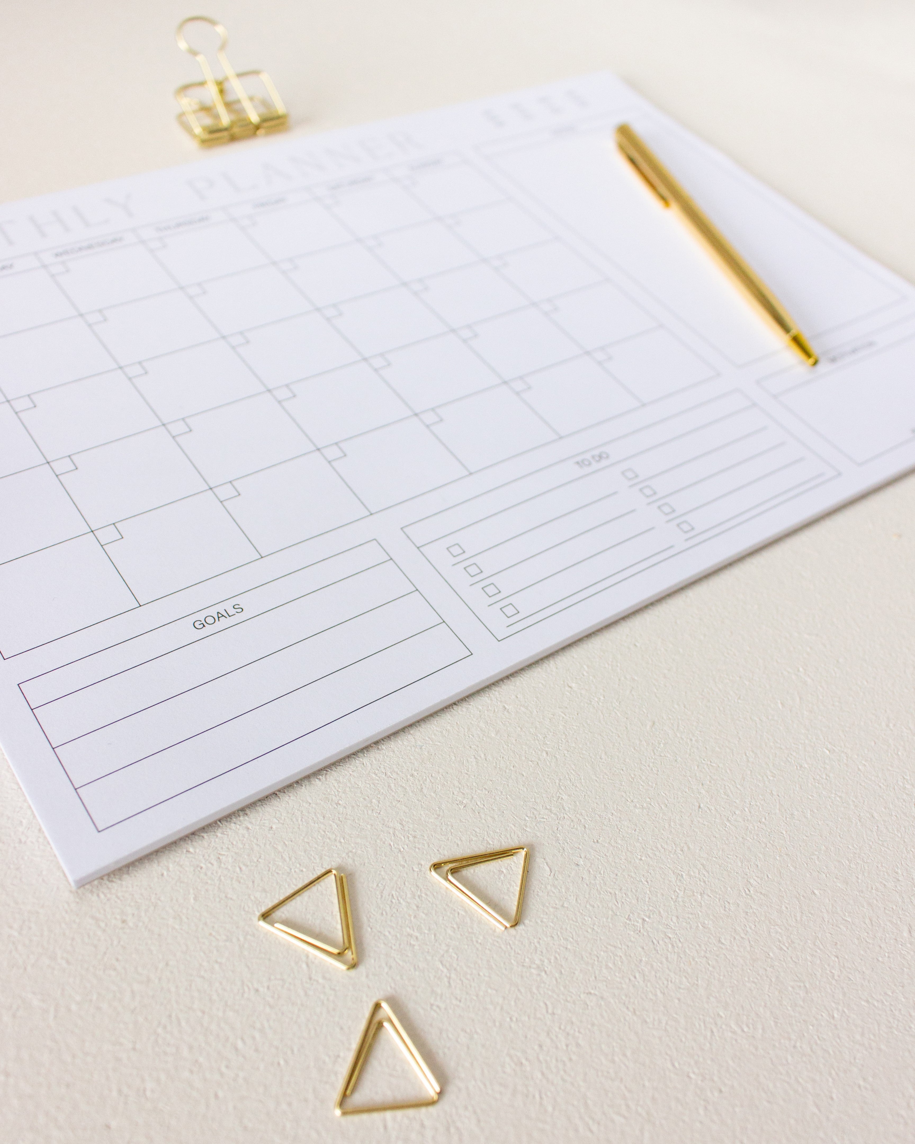 Undatierter Monatsplaner DIN A4 Schreibtischunterlage mit goldenem Kugelschreiber, goldener Foldbackklammer und goldenen dreieckigen Büroklammern auf hell-beigem Untergrund.